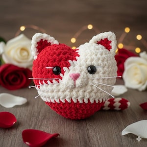 May include: A handmade crochet cat with a red and white color scheme. The cat has black eyes, pink nose, and white whiskers. The cat is sitting on a wooden surface with rose petals and roses in the background.