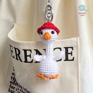 Puede incluir: Llavero de ganso blanco de crochet con un sombrero rojo. El ganso tiene patas naranjas y un pico amarillo. El llavero está unido a un llavero plateado.