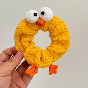 May include: A yellow crocheted hair scrunchie shaped like a cartoon chick. The scrunchie has white eyes with black pupils, an orange beak, and orange feet. The hair accessory is handmade and suitable for all ages.