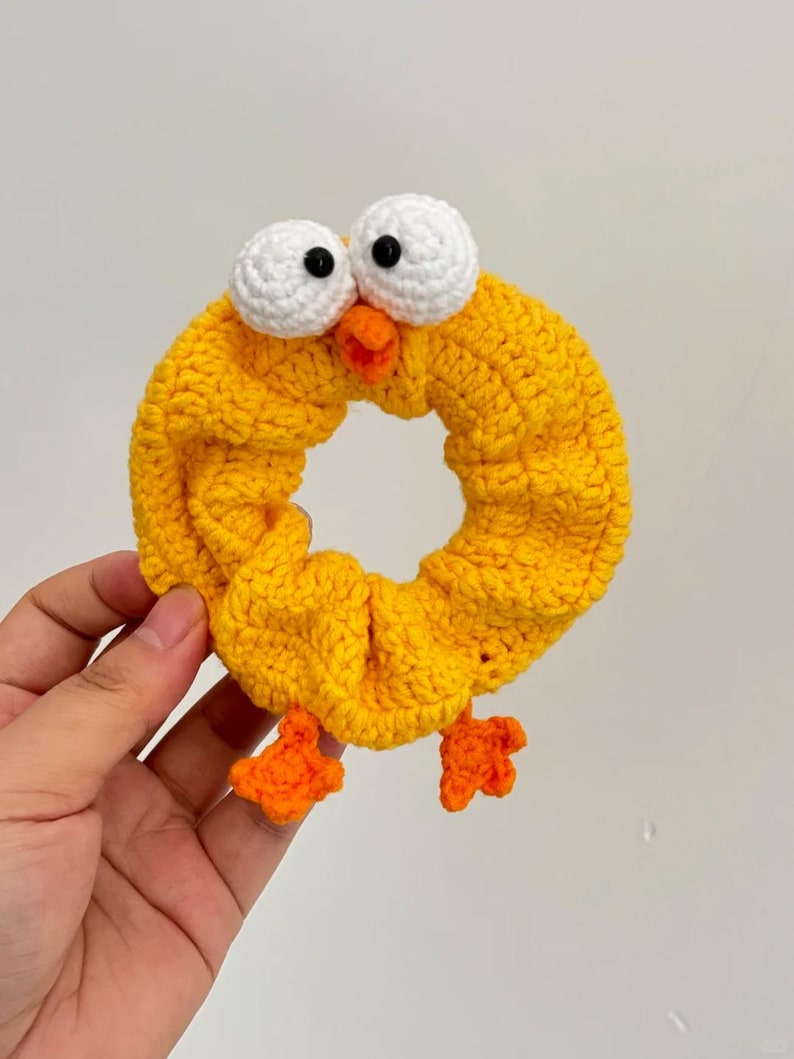 May include: A yellow crocheted hair scrunchie shaped like a cartoon chick. The scrunchie has white eyes with black pupils, an orange beak, and orange feet. The hair accessory is handmade and suitable for all ages.