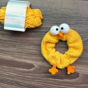 May include: A yellow crochet scrunchie in the shape of a chick with white eyes, an orange beak, and orange feet. A ball of yellow yarn with a label is next to the scrunchie. The items are on a wooden surface.