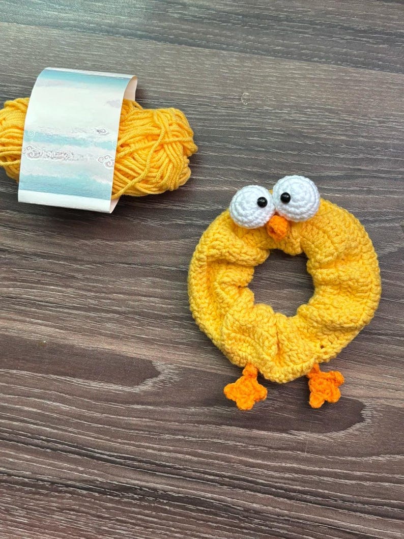 May include: A yellow crochet scrunchie in the shape of a chick with white eyes, an orange beak, and orange feet. A ball of yellow yarn with a label is next to the scrunchie. The items are on a wooden surface.