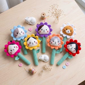 May include: A collection of seven crocheted flower rattles, each featuring a cat face at the center. The flowers have petals in various colors including blue, yellow, purple, orange, white, red, and pink, all attached to green stems.