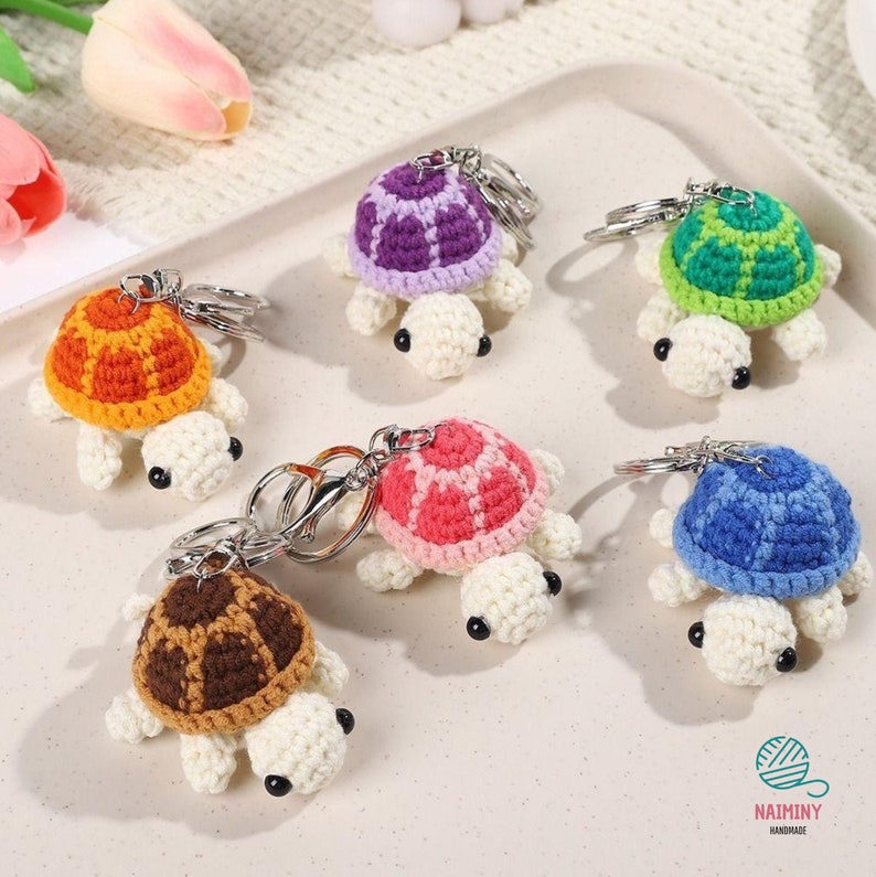 Op de afbeelding: Zes gehaakte schildpad sleutelhangers in verschillende kleuren: oranje, paars, groen, roze, blauw en bruin. Elke schildpad heeft een wit lichaam en een zilveren sleutelring.