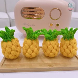 Könnte beinhalten: Vier gehäkelte Ananas-Ornamente in Gelb und Grün. Die Ananas liegen auf einer Holzoberfläche.