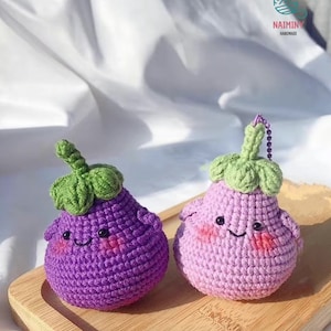 Puede incluir: Dos llaveros de berenjena hechos a mano con ganchillo. Uno es morado, el otro rosa. Ambos tienen copas verdes con hojas, caras sonrientes y mejillas rosadas. Están sobre una bandeja de madera. De NAIMINY HANDMADE.