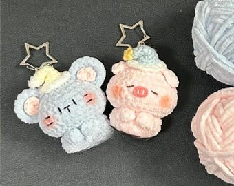 Puede incluir: Dos llaveros de peluche: un ratón azul y un cerdo rosa, cada uno con un pequeño sombrero y sujeto a un llavero plateado en forma de estrella. El ratón tiene las letras "IT" bordadas en su cuerpo. Bolas de hilo en azul claro y rosa en el fondo.