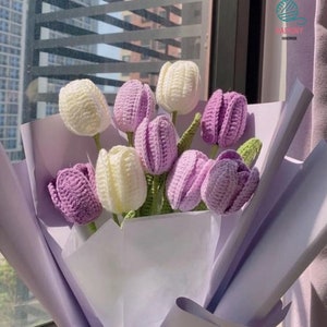 Może przedstawiać: Bukiet dzianych tulipanów w kolorze fioletowym i białym, owinięty w jasnofioletowy papier tiulowy. Bukiet jest oznaczony etykietą "Raiminy Handmade".