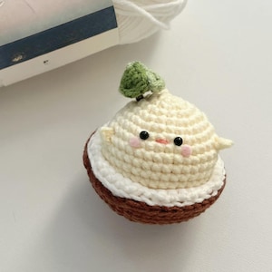 Pode incluir: Um brinquedo de amigurumi de crochê em forma de coco com uma folha verde no topo. O coco tem uma casca branca e uma base marrom. O brinquedo tem um rosto bonito com olhos pretos e bochechas rosadas.