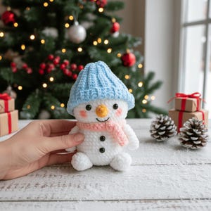 May include: A crocheted snowman figure with a light blue hat and a pink scarf. The snowman has a carrot nose, black button eyes, and rosy cheeks. A festive decoration for the winter holidays.