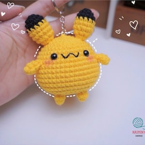 Pode incluir: Um chaveiro de crochê amarelo com orelhas pretas e um rosto bonito. O chaveiro tem a forma de um animal pequeno com um corpo redondo e pernas curtas.
