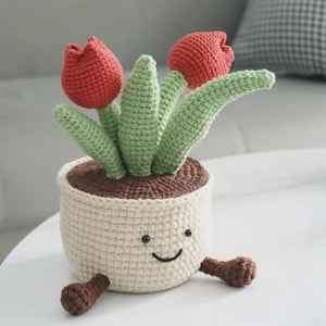 May include: A crocheted potted plant featuring two red tulip blossoms and green leaves emerging from a cream-colored pot. The pot has a stitched smiling face with two black eyes and a curved mouth, and two small brown feet extending from the base.