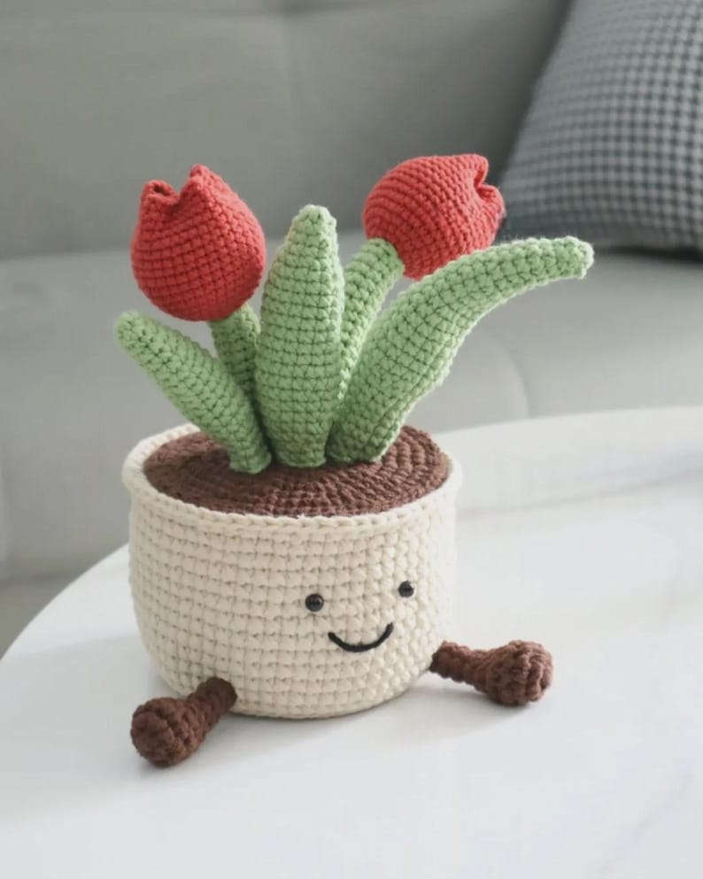 May include: A crocheted potted plant featuring two red tulip blossoms and green leaves emerging from a cream-colored pot. The pot has a stitched smiling face with two black eyes and a curved mouth, and two small brown feet extending from the base.