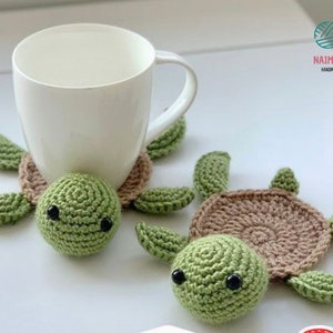 May include: Two crocheted turtle coasters in green and brown. The coasters are shaped like turtles with a round brown shell and green heads and legs. A white mug is sitting on one of the coasters.