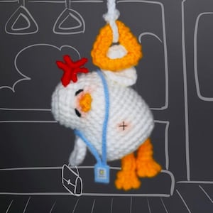 Pode incluir: Uma pelúcia de galinha em crochê branca com crista vermelha, bico laranja e pés laranja. Pendura-se num anel triangular amarelo por um cordão branco. A galinha usa um cordão azul claro com uma pequena etiqueta retangular e um ponto de cruz.