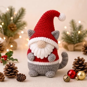 Pode incluir: Uma figura de gato gnomo de crochê usando um gorro de Papai Noel vermelho com um pompom branco, uma barba branca e um suéter vermelho e branco. O gato tem orelhas cinzas, corpo cinza e cauda cinza, com um pequeno nariz bege acima da barba.