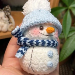 May include: A handcrafted snowman figure wearing a light blue knitted hat and scarf. The snowman has an orange carrot nose, pink cheeks, and a white pom-pom on the hat. Two blue buttons are on the snowman's body.