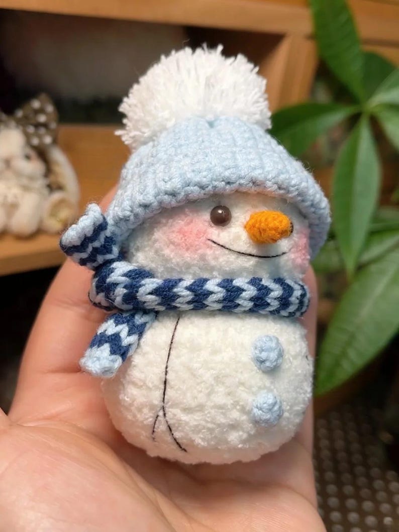 May include: A handcrafted snowman figure wearing a light blue knitted hat and scarf. The snowman has an orange carrot nose, pink cheeks, and a white pom-pom on the hat. Two blue buttons are on the snowman's body.