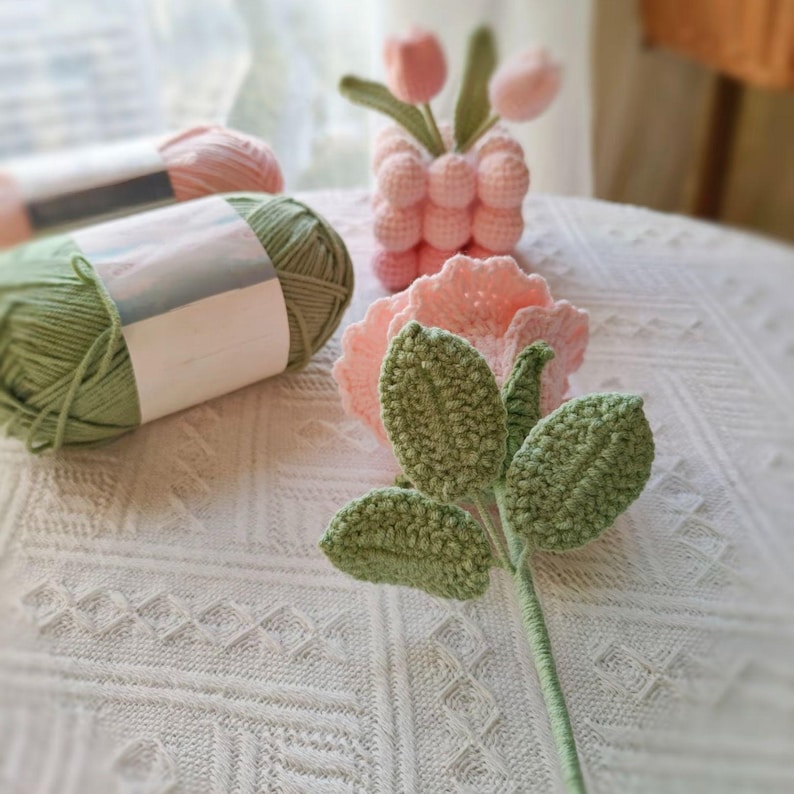 May include: A crocheted pink rose with green leaves and a stem. The rose is sitting on a white crocheted surface with two balls of yarn, one green and one pink.