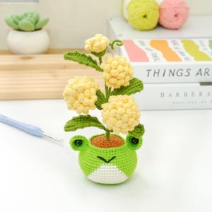 May include: A handmade crochet flower arrangement featuring yellow pom-pom flowers, green leaves, and a frog-shaped pot. The pot is green and white, with black eyes. The arrangement is on a white surface.