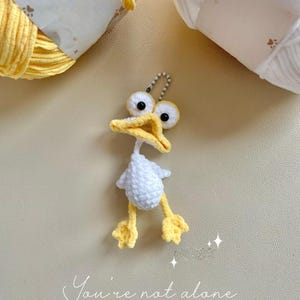 May include: A handmade crochet duck keyring with a white body, yellow feet, and a yellow beak. The duck has large, googly eyes and a silver keyring attachment. The text "You're not alone" is written in cursive below the duck.