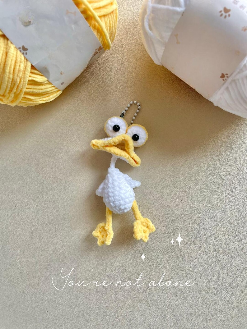 May include: A handmade crochet duck keyring with a white body, yellow feet, and a yellow beak. The duck has large, googly eyes and a silver keyring attachment. The text "You're not alone" is written in cursive below the duck.
