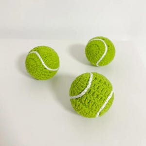 Op de afbeelding: Drie handgemaakte, groene gehaakte tennisballen met witte stikseldetails. De ballen zijn geplaatst op een wit oppervlak, wat de textuur en het vakmanschap van het garen benadrukt. Een speels en uniek item.