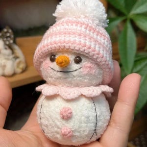 May include: A handmade crochet snowman wearing a pink and white striped hat with a white pom-pom. The snowman has an orange carrot nose, rosy cheeks, and a pink scalloped collar. Two pink buttons are on the body. Black thread outlines the body.
