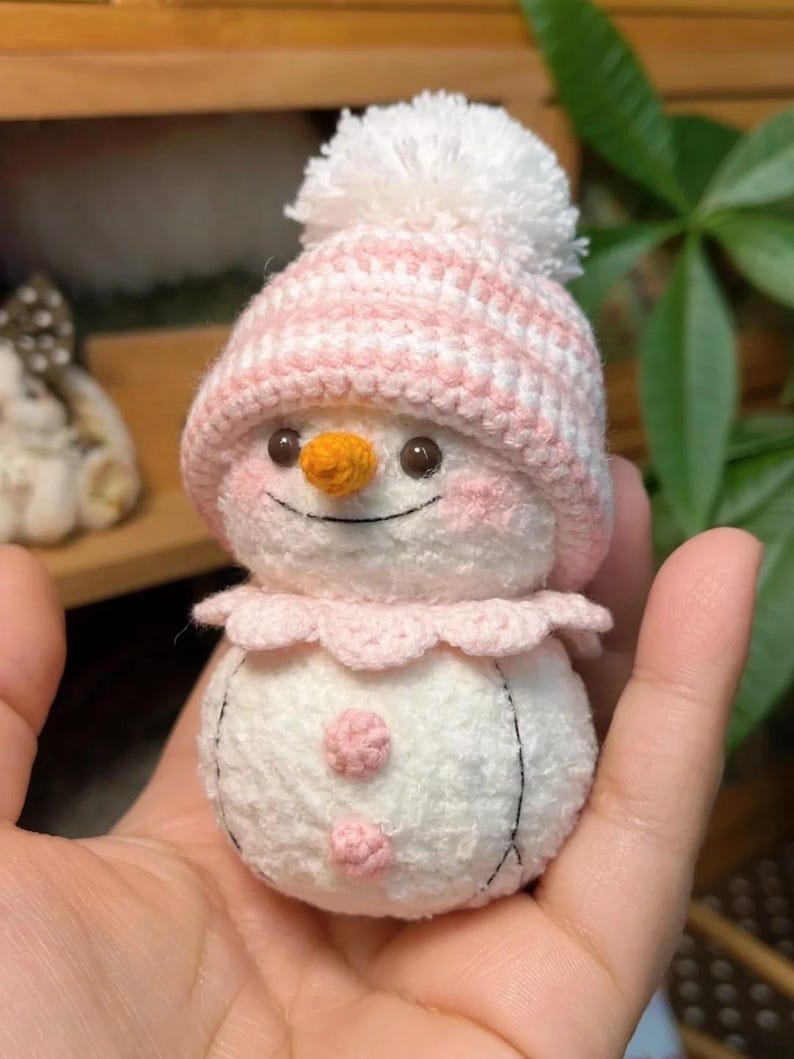 May include: A handmade crochet snowman wearing a pink and white striped hat with a white pom-pom. The snowman has an orange carrot nose, rosy cheeks, and a pink scalloped collar. Two pink buttons are on the body. Black thread outlines the body.