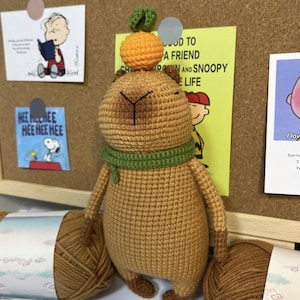 Op de afbeelding: Een handgemaakte gehaakte capybarapop in bruintinten, met een groene sjaal en bekroond met een oranje en groene gehaakte vrucht. De pop houdt twee bollen garen vast. Achtergrond bevat Peanuts-kunstwerken en een prikbord.