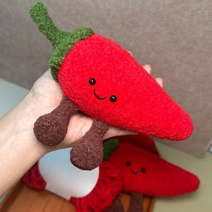 Puede incluir: Dos pimientos rojos de peluche con caras sonrientes y patas marrones. Uno se sostiene en alto, el otro descansa debajo. Los pimientos tienen tallos verdes y están hechos de un material suave y texturizado.