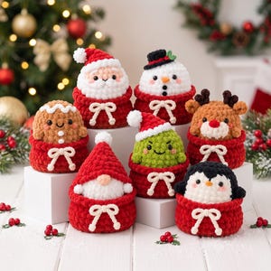 Puede incluir: Una colección de siete figuras de peluche tejidas a ganchillo que incluyen a Santa Claus, un muñeco de nieve, un hombre de jengibre, un reno, un gnomo, una criatura verde y un pingüino, cada uno en una bolsa roja con un lazo blanco.