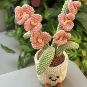 Puede incluir: Una maceta de flores de ganchillo con una cara sonriente, patas marrones y flores de color rosa claro con centros amarillos. La maceta es de color crema y contiene hojas y tallos verdes. El art&iacute;culo hecho a mano es una pieza decorativa caprichosa.