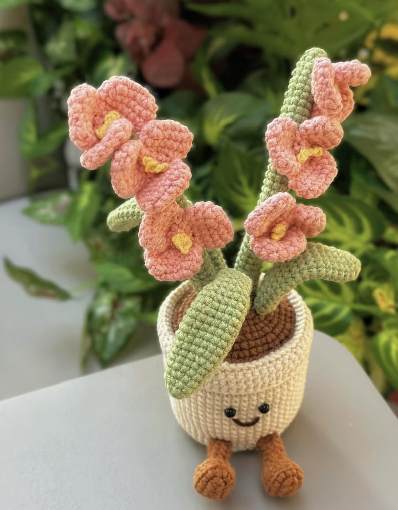 Puede incluir: Una maceta de flores de ganchillo con una cara sonriente, patas marrones y flores de color rosa claro con centros amarillos. La maceta es de color crema y contiene hojas y tallos verdes. El art&iacute;culo hecho a mano es una pieza decorativa caprichosa.