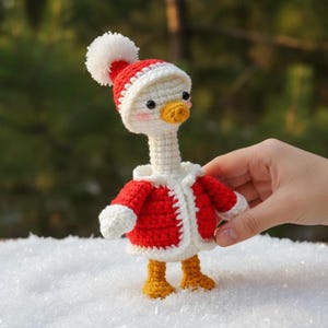 May include: A handmade crochet goose wearing a red and white Santa hat and jacket. The goose has a white head and neck, a yellow beak and feet, and is standing in the snow. The goose is a festive holiday decoration.