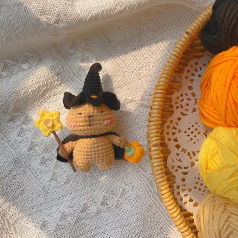 May include: A handmade crochet Halloween-themed figure of a bear wearing a witch hat and cape, holding a star wand and a pumpkin. The bear is tan with black accents. The background includes a basket with yarn.