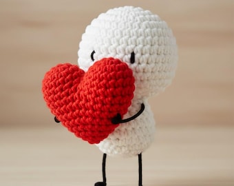 May include: A handmade white crochet figure holding a red heart. The figure has black eyes, arms, and legs. The heart is a vibrant red color, and the figure is standing upright.