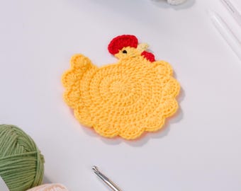 May include: A handmade, yellow crochet chicken coaster with a red comb and wattle. The coaster is round with a scalloped edge. A crochet hook and yarn are also visible.
