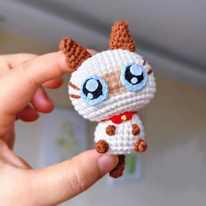 May include: A crocheted cat figure with a white body, brown ears, and large blue eyes. It has a red collar with a yellow button. The cat's paws and tail are brown. A handmade item.