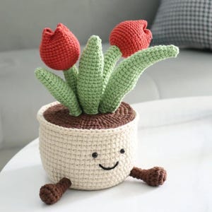 May include: A crocheted plant pot with a smiling face, holding two red tulip flowers with green leaves. The pot is cream-colored with brown soil and small brown feet.