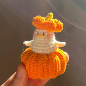 May include: A handmade crochet pumpkin-shaped figure with a white body and orange accents. The figure has a small, round head with a yellow stem and is wearing a pumpkin hat. The figure is held in a hand.