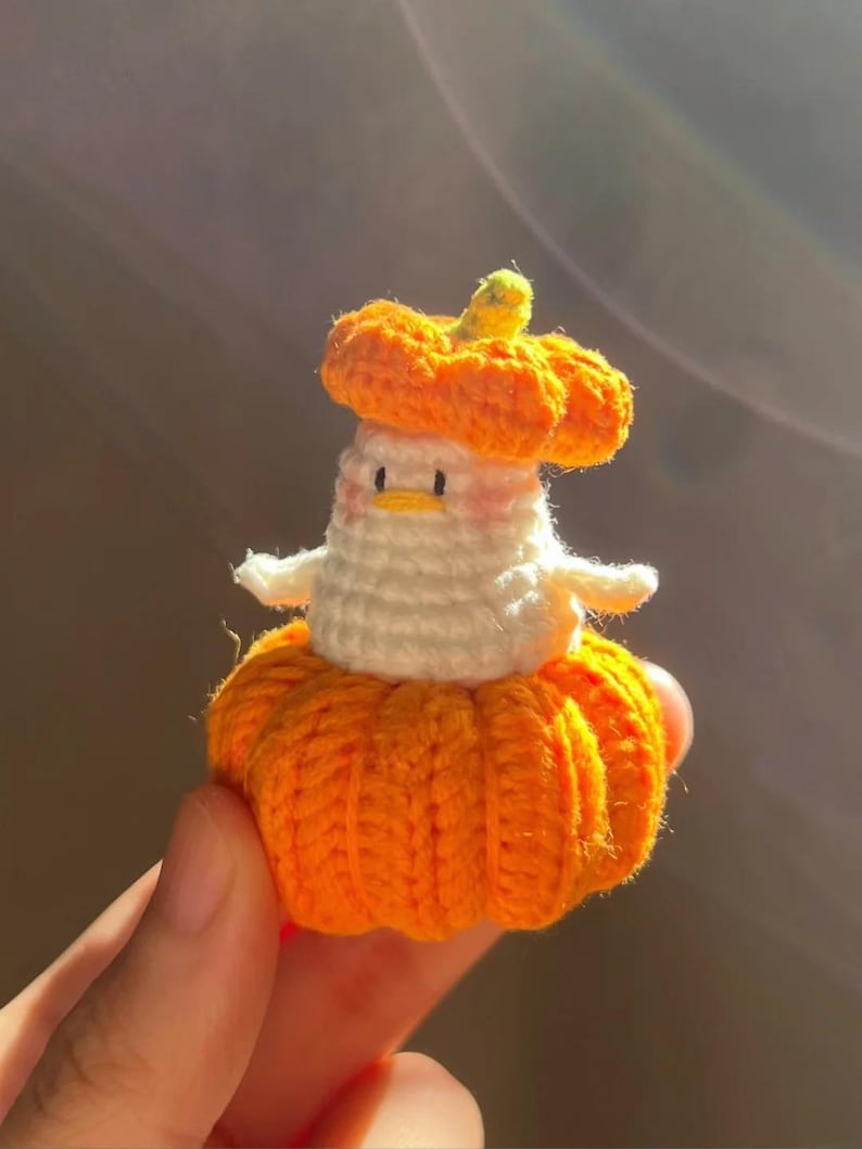 May include: A handmade crochet pumpkin-shaped figure with a white body and orange accents. The figure has a small, round head with a yellow stem and is wearing a pumpkin hat. The figure is held in a hand.