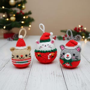 May include: Three handmade crochet Christmas ornaments shaped like animals, each wearing a Santa hat. The ornaments are beige, white, and gray, with red and green accents. They have embroidered details and hanging loops.