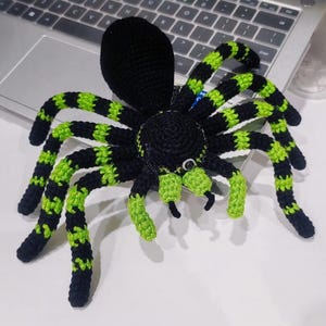 May include: A handmade crochet spider in black and neon green. The spider has a large, round body and eight long legs with alternating stripes of black and green. The spider is sitting on a white surface near a laptop.