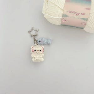 K&ouml;nnte beinhalten: Ein handgefertigter H&auml;kel-Schl&uuml;sselanh&auml;nger mit einer wei&szlig;en Katze und einem kleinen blauen Fisch-Anh&auml;nger. Die Katze hat rosa Akzente und einen sternf&ouml;rmigen Verschluss. Ein Kn&auml;uel wei&szlig;e Wolle befindet sich im Hintergrund.
