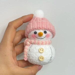 May include: A handmade snowman figurine with a pink knitted hat and scarf. The snowman has a white body, black eyes, rosy cheeks, and an orange carrot nose. Two wooden buttons are sewn on the front.