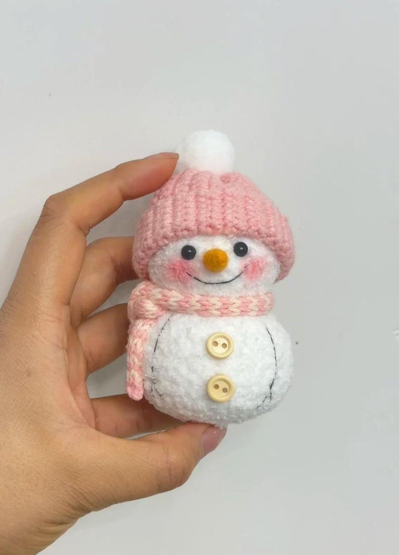 May include: A handmade snowman figurine with a pink knitted hat and scarf. The snowman has a white body, black eyes, rosy cheeks, and an orange carrot nose. Two wooden buttons are sewn on the front.