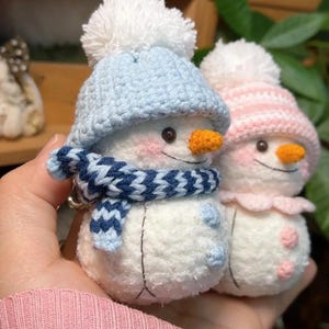 May include: Two handmade snowman keyrings. One snowman wears a blue hat and scarf, the other a pink hat. Both have orange carrot noses, rosy cheeks, and black button eyes. The snowmen are made of white yarn.