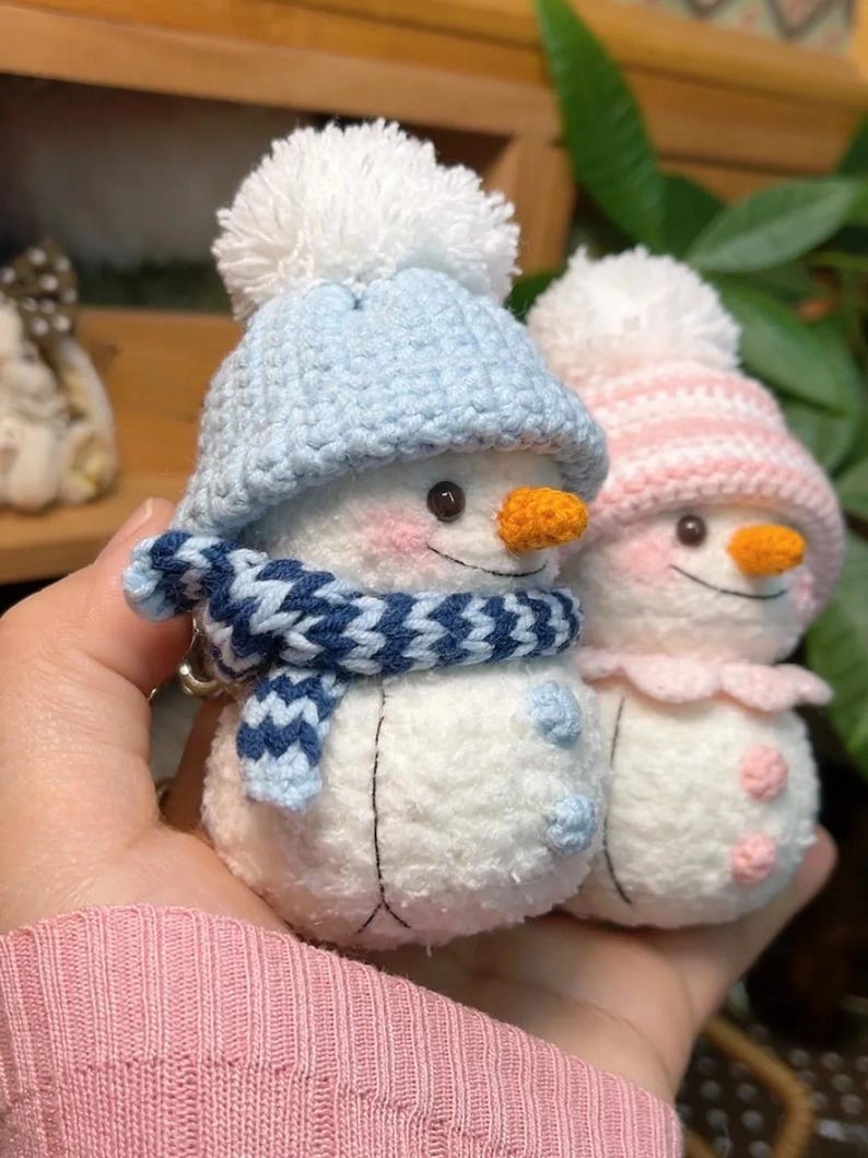 May include: Two handmade snowman keyrings. One snowman wears a blue hat and scarf, the other a pink hat. Both have orange carrot noses, rosy cheeks, and black button eyes. The snowmen are made of white yarn.