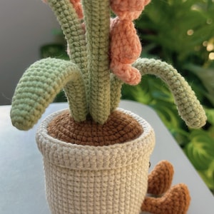 Puede incluir: Planta de ganchillo hecha a mano en una maceta blanca con tierra marr&oacute;n. La planta tiene tallos y hojas verdes y flores rosas. La maceta tiene peque&ntilde;os pies marrones. El dise&ntilde;o es caprichoso.